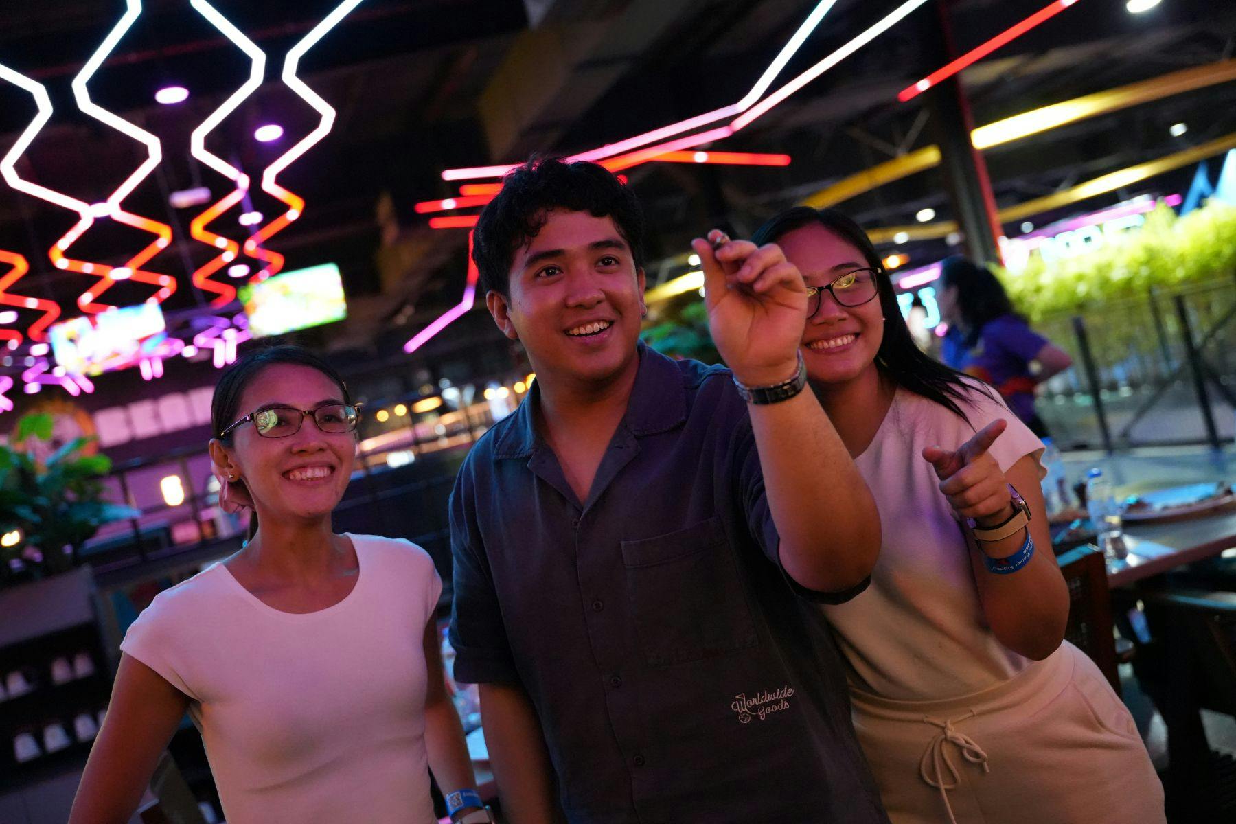 Τρεις άνθρωποι χαμογελούν και παίζουν βελάκια σε έναν χώρο με πολύχρωμα φώτα νέον πάνω από το κεφάλι τους.