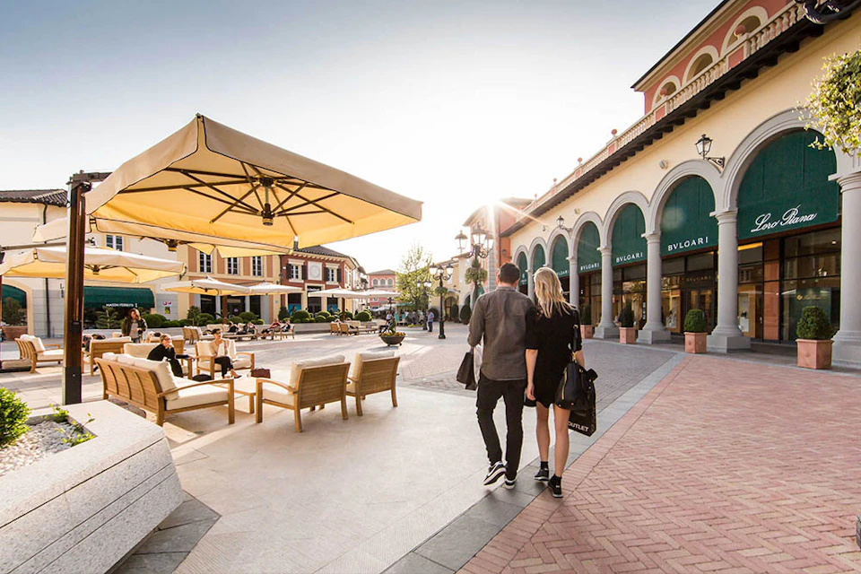 A couple walks through an outdoor shopping area with stores, seating, and large umbrellas under a clear sky.