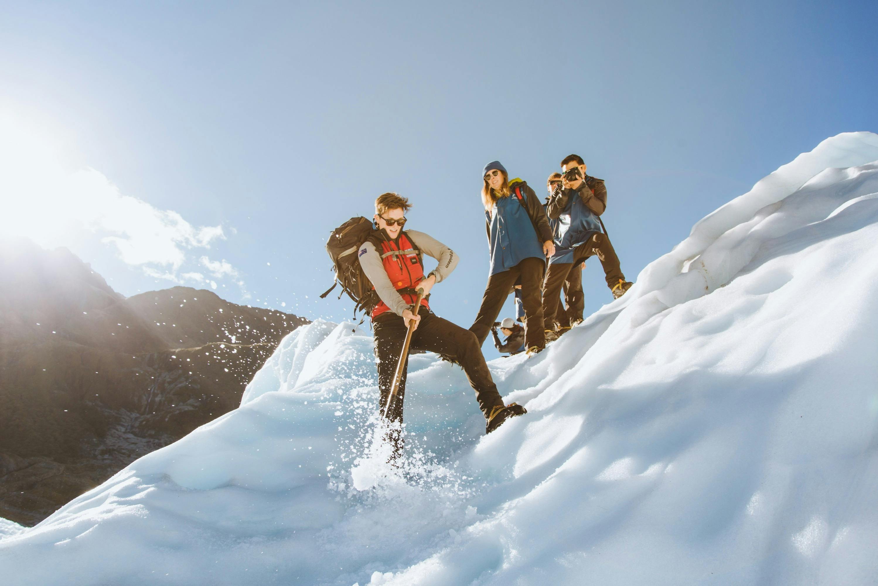 Three people hiking on a snowy slope, one using an ice axe while others observe and take photos under clear skies.