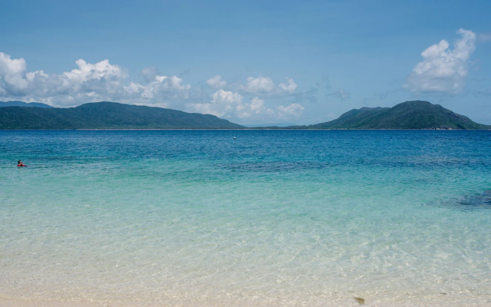 A calm beach with clear blue water, green hills in the distance, and a bright, partly cloudy sky.