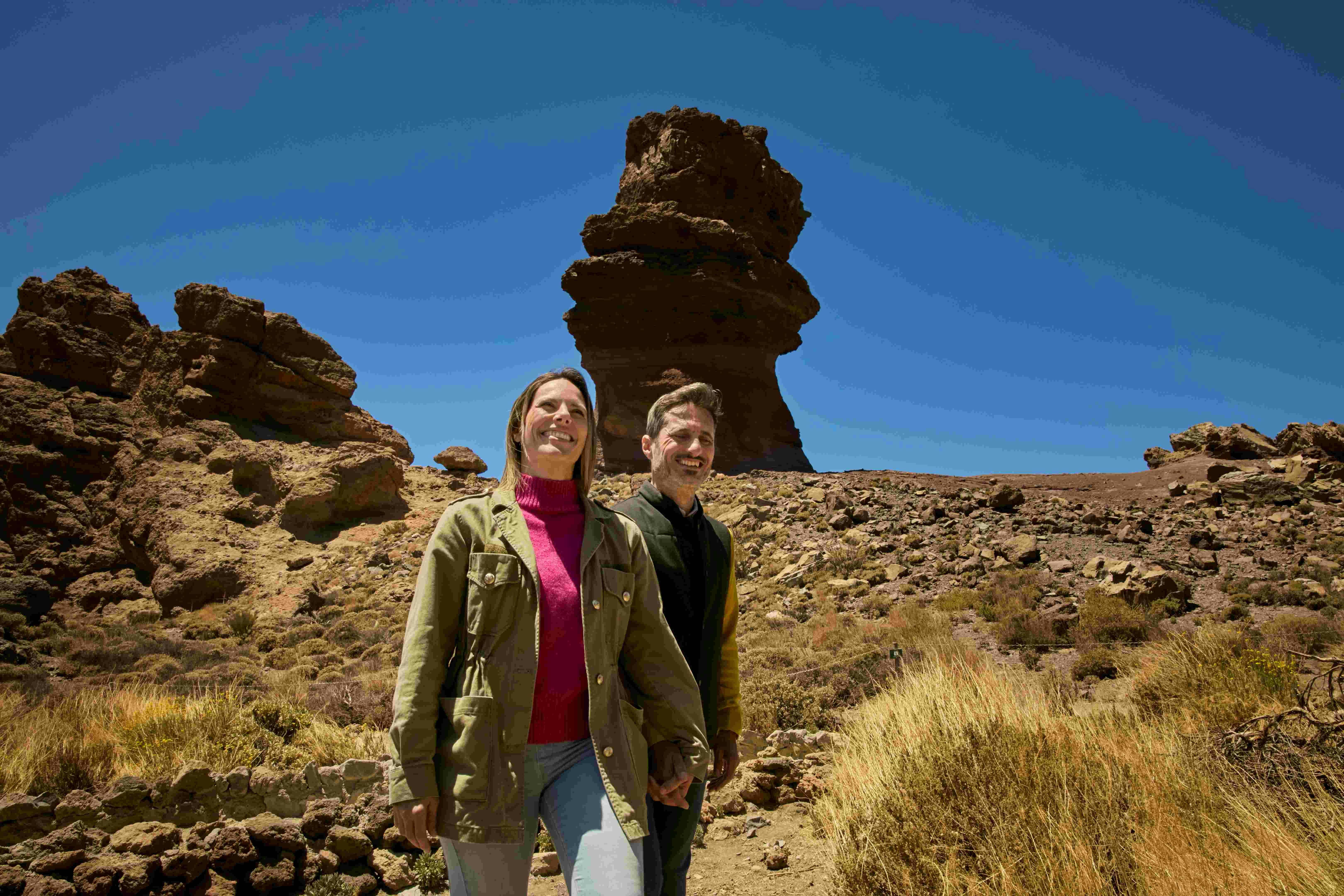 A smiling woman and man stand together in a rocky desert landscape with a distinctive tall rock formation in the background.