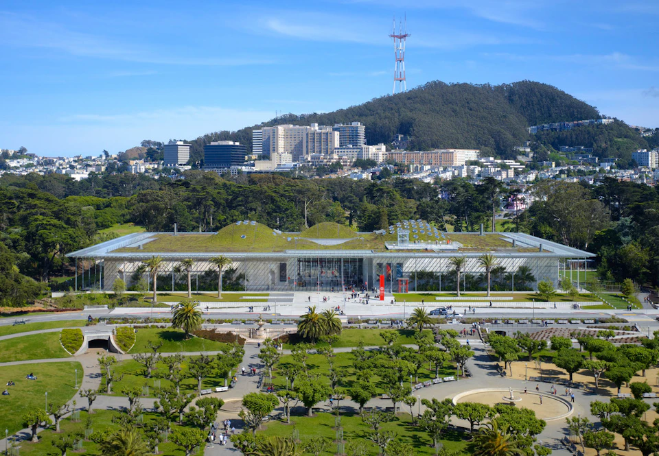 A large building with a green roof is surrounded by a park with many people. Hills, tall buildings, and a tower are in the background.