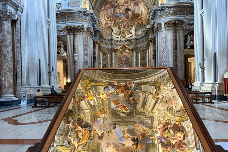 A detailed ceiling fresco in a richly decorated church, with a mirror on a stand reflecting the artwork below.
