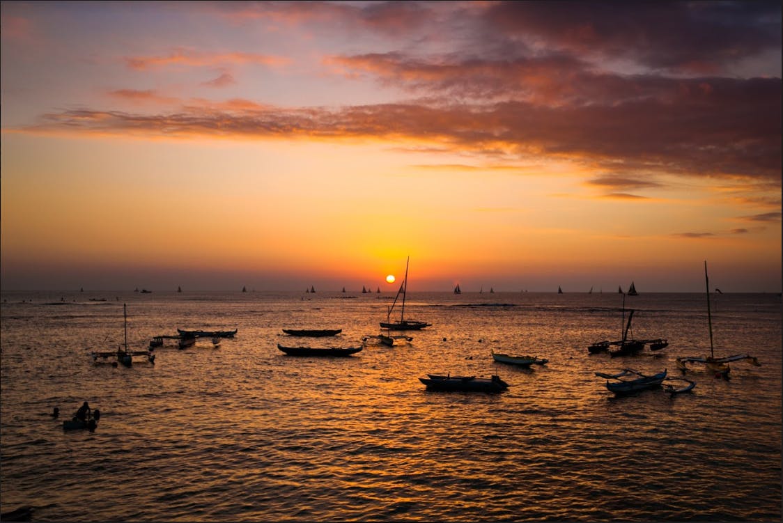 Sailboats and rowboats dot a calm sea at sunset, with the sun low on the horizon and a sky streaked with orange and purple clouds.