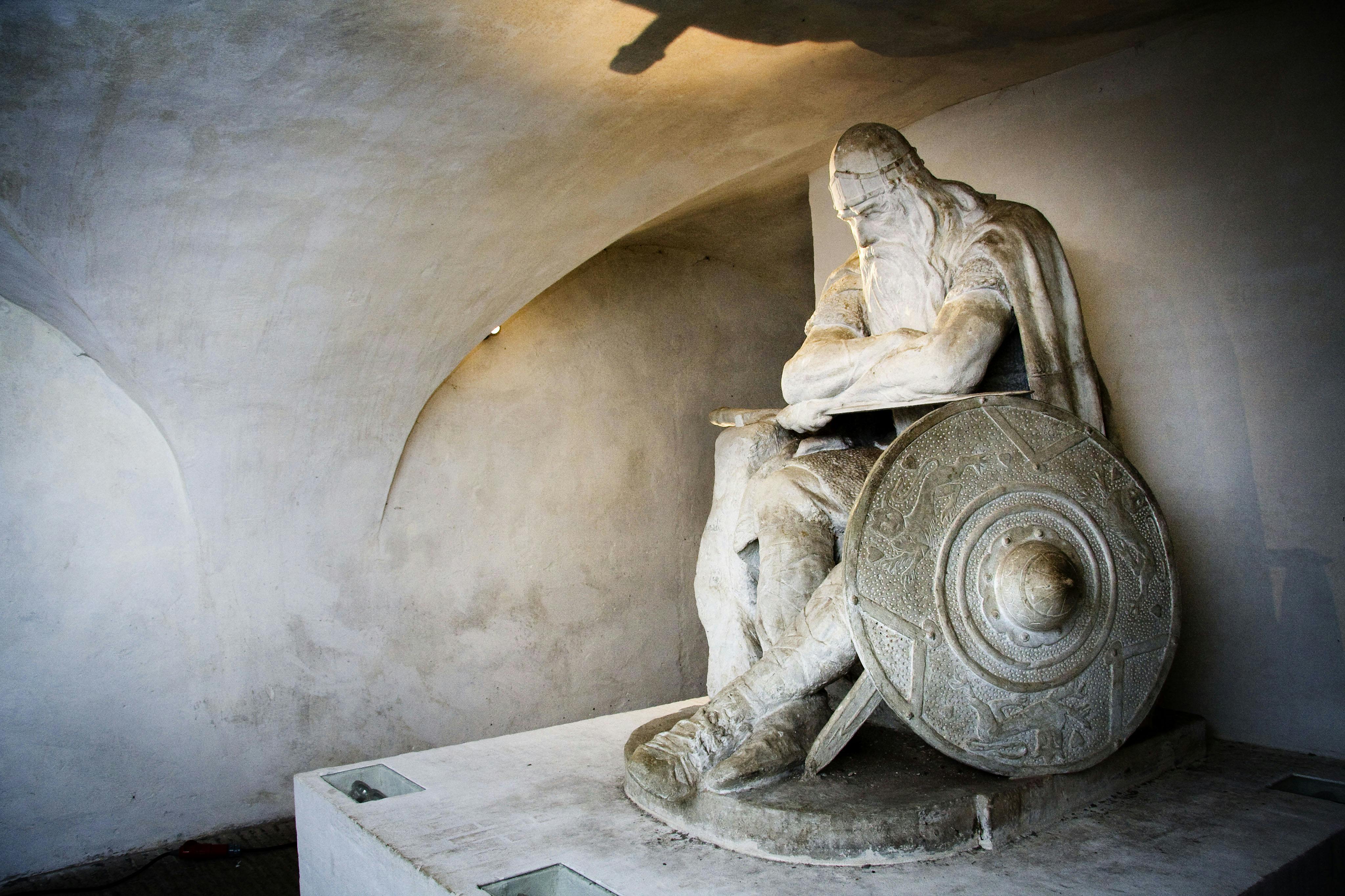 Stone statue of a seated, bearded man with a shield and staff, set against an arched, minimalist background.