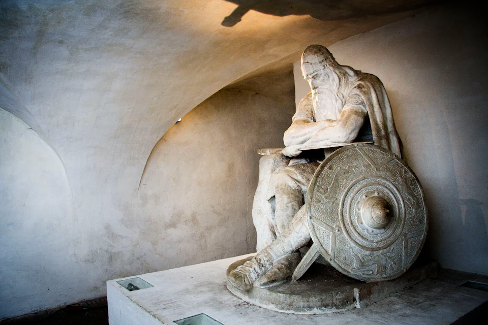 Stone statue of a seated, bearded man with a shield and staff, set against an arched, minimalist background.