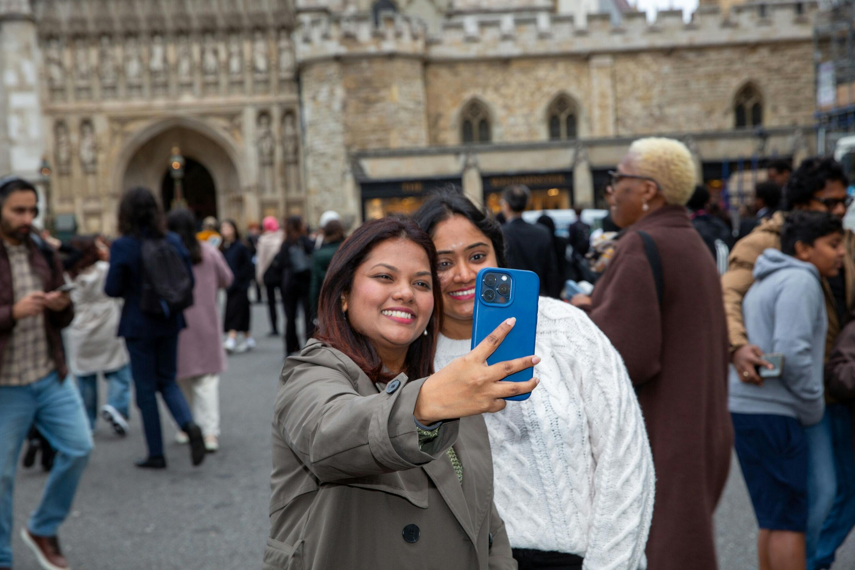 两人在一栋历史悠久的建筑前摆姿势自拍，背景是一群人。