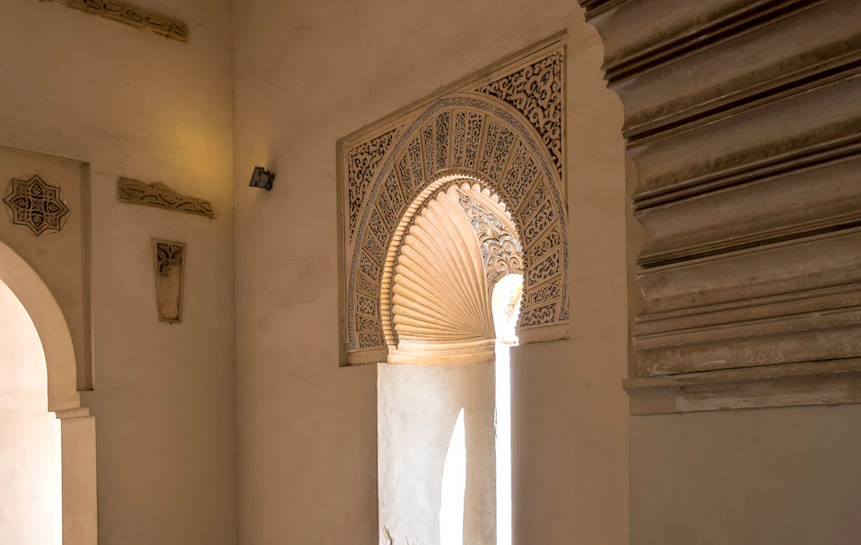 Ornate arched window with intricate carvings and a fan-shaped architectural detail, casting light into a minimalist room.