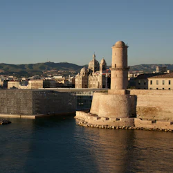 Ett historiskt fort intill en modern byggnad vid vattnet, med en stadsbild och berg i bakgrunden under en klar himmel.