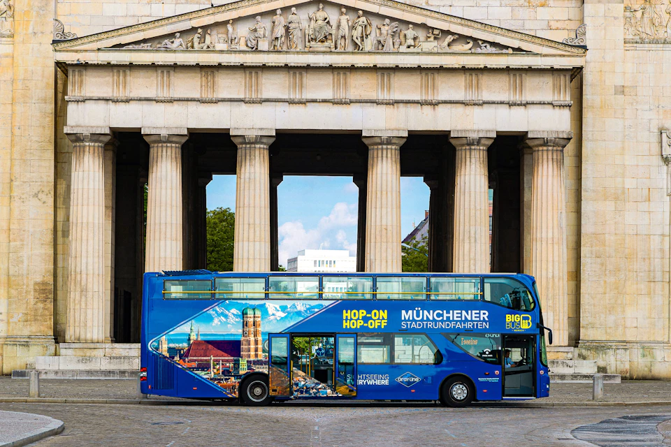 A blue double-decker tour bus labeled 'Hop-On Hop-Off Münchener Stadtrundfahrten' in front of a historic colonnade.