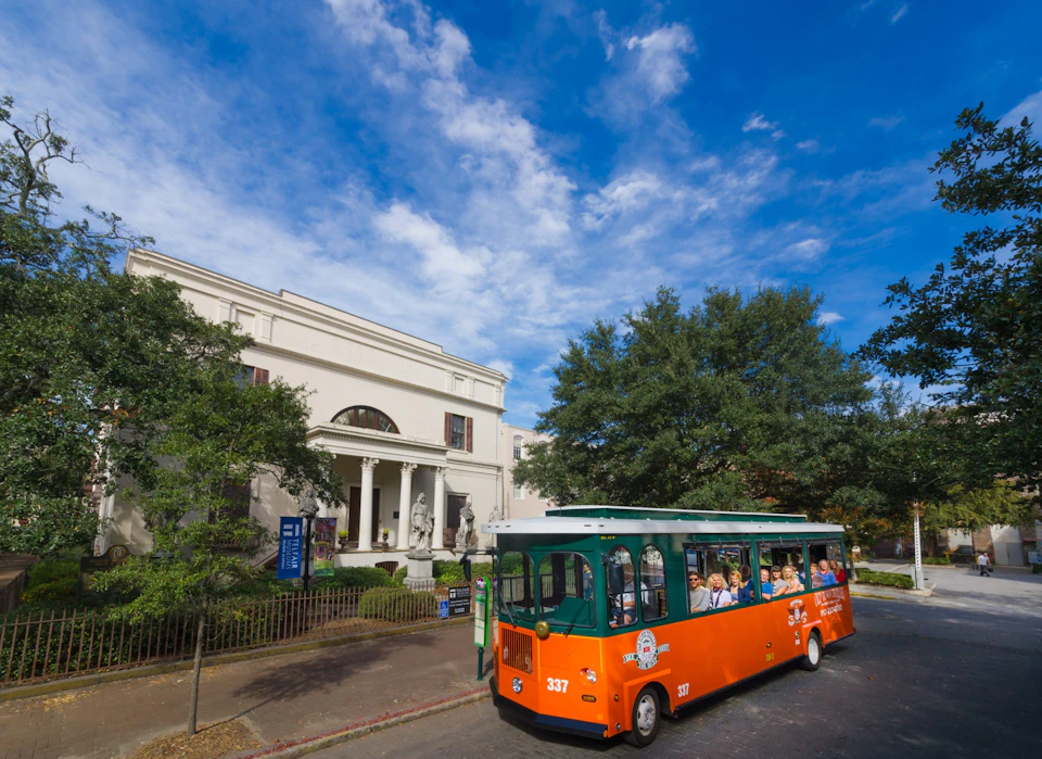 An orange and green trolley with passengers passes by a large white building with columns and statues, under a partly cloudy sky.