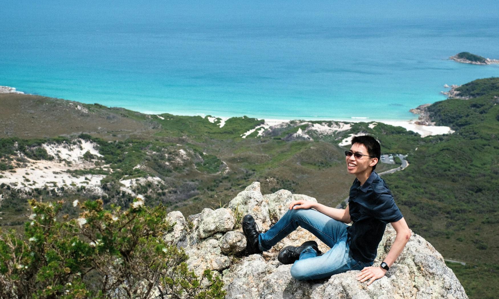 Άτομο που κάθεται σε βραχώδη βράχο, χαμογελαστό, με καταπράσινο τοπίο και τιρκουάζ ωκεανό στο παρασκήνιο.