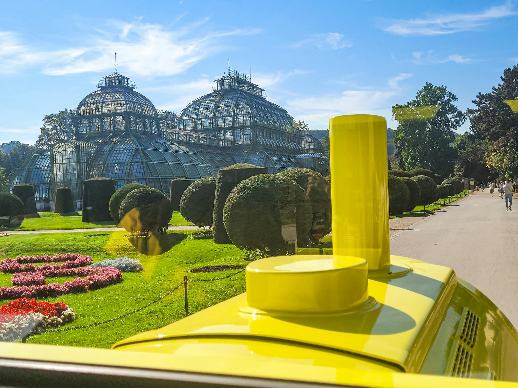 Gult tåg framför en stor vinterträdgård i glas med kupolformade tak, omgiven av välskötta trädgårdar och träd.