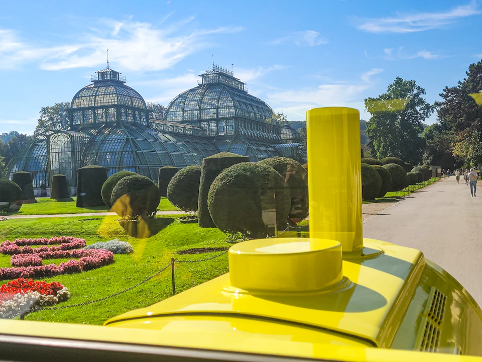 Yellow train in front of a large glass conservatory with dome-shaped roofs, surrounded by manicured gardens and trees.