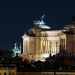 Upplyst neoklassisk byggnad på natten med pelare och statyer av vagnar, sedda på avstånd mot en mörk himmel.