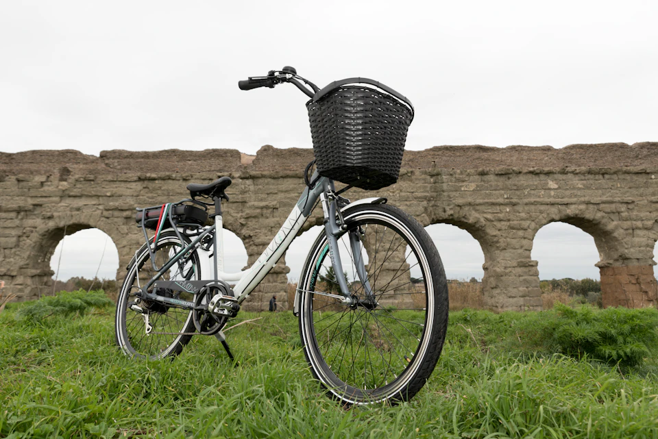 A bicycle with a front basket is parked on grass in front of a stone aqueduct on an overcast day.