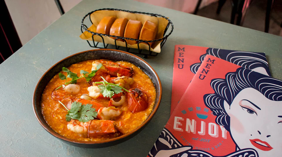 A bowl of seafood stew garnished with herbs, a basket of bread rolls, and menus on a green table.