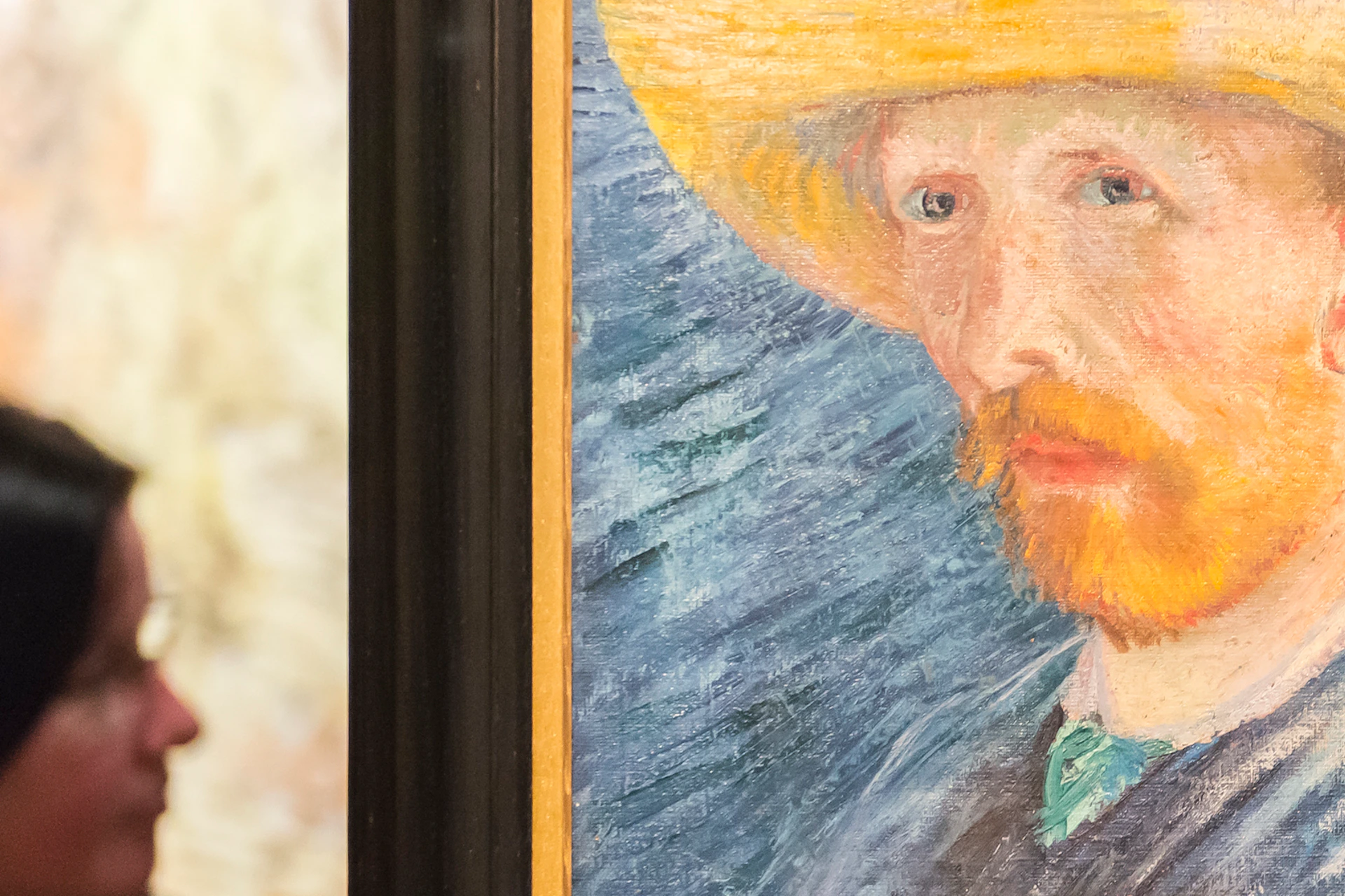 A framed painting of a man with a yellow hat and a red beard, displayed in a gallery with two people talking in the background.
