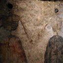 Due figure scheletriche che si tengono per mano, dipinte su un muro di pietra antico e strutturato. Le figure sono prive di testa e indossano abiti colorati.
