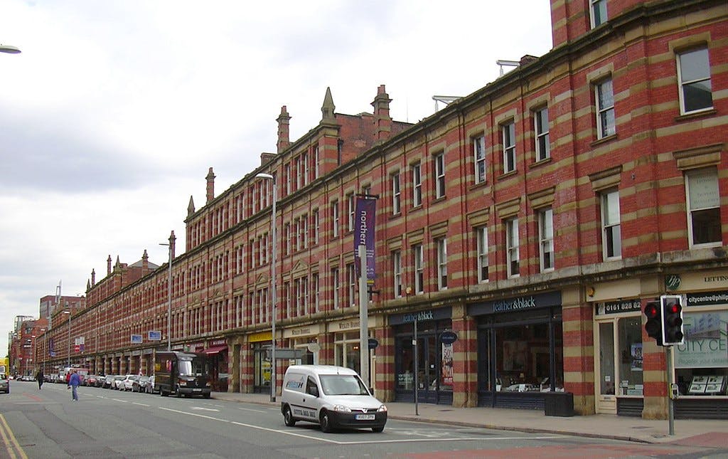 Ulice lemovaná červenými cihlovými budovami s obchody, auty zaparkovanými podél silnice a dodávkou jedoucí po silnici.