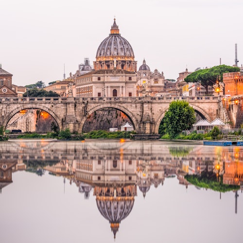 Il ponte storico e la cupola della Basilica di San Pietro si riflettono in un fiume tranquillo, illuminato dalle luci della sera.