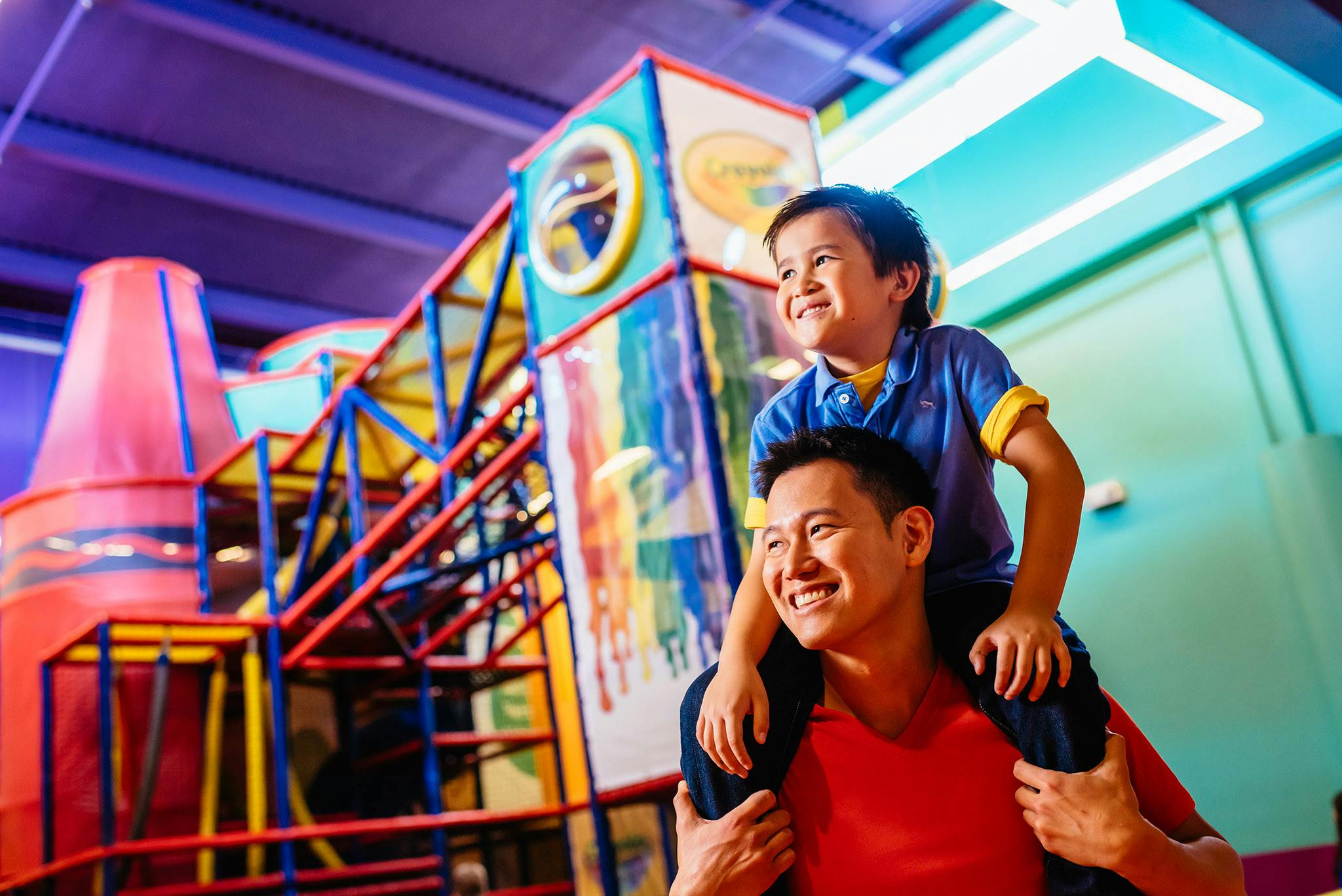 Smiling man carrying a boy on his shoulders inside a colorful indoor play area with climbing structures.