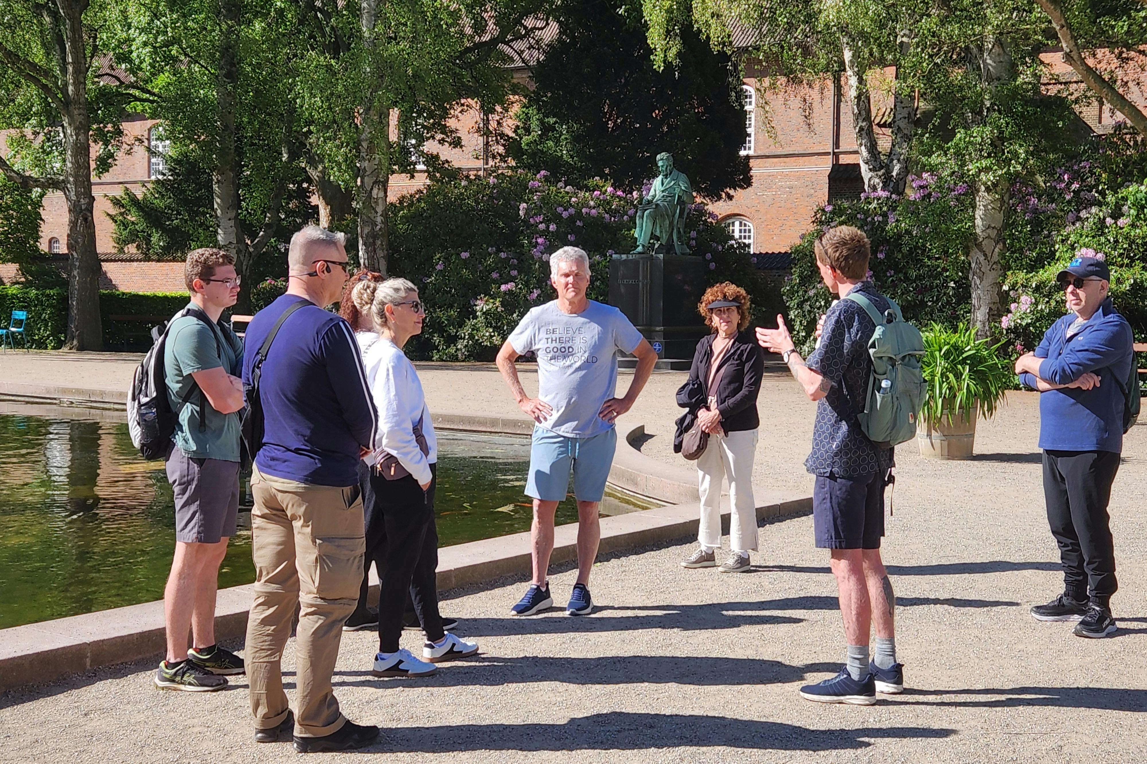 A group of people standing near a pond in a park with trees, a statue, and a building in the background.