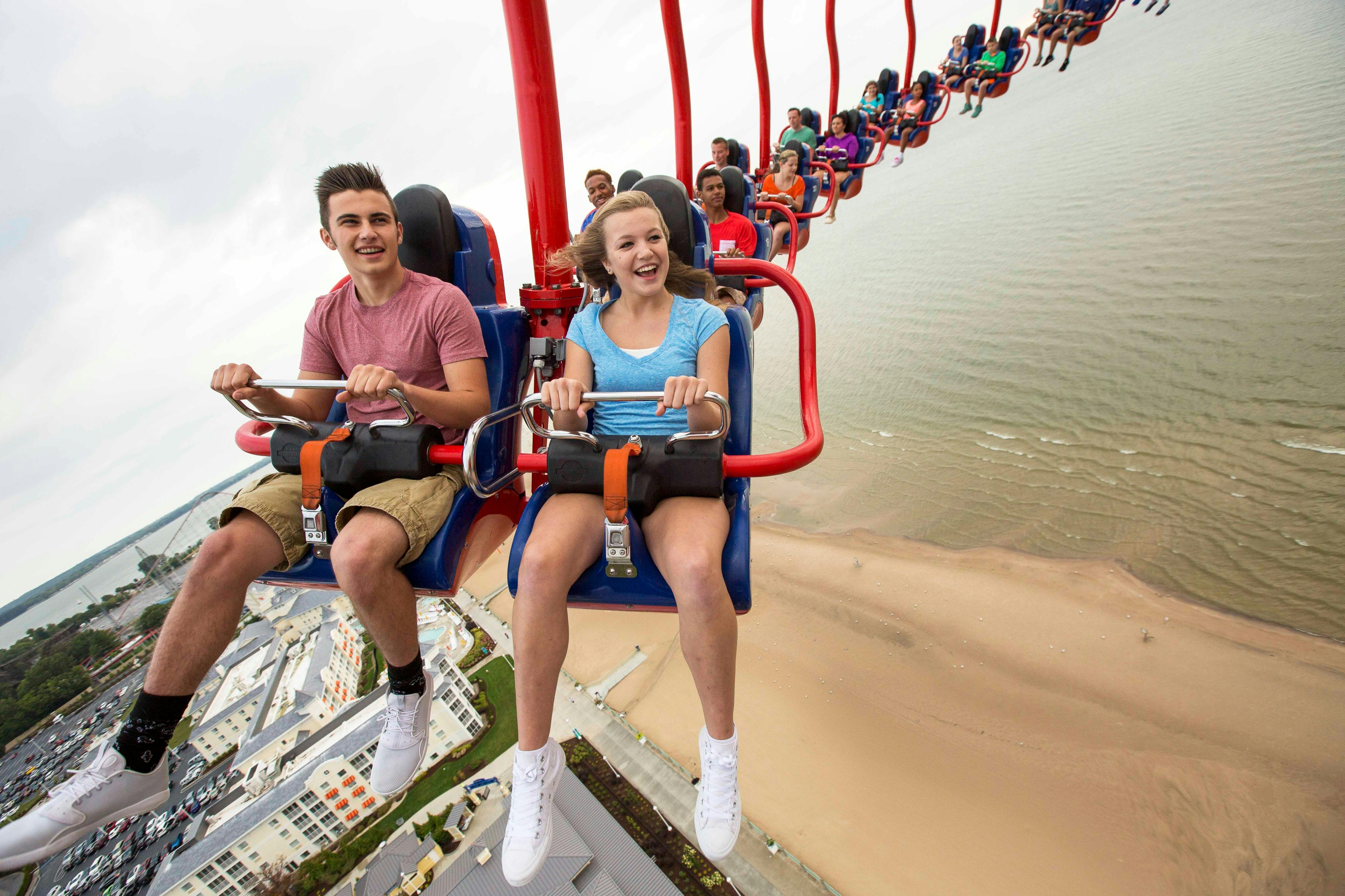 Άνθρωποι που απολαμβάνουν μια βόλτα με ψηλά πόδια με θέα μια παραλία και κτίρια, χαμογελώντας και κρατώντας τις μπάρες ασφαλείας.