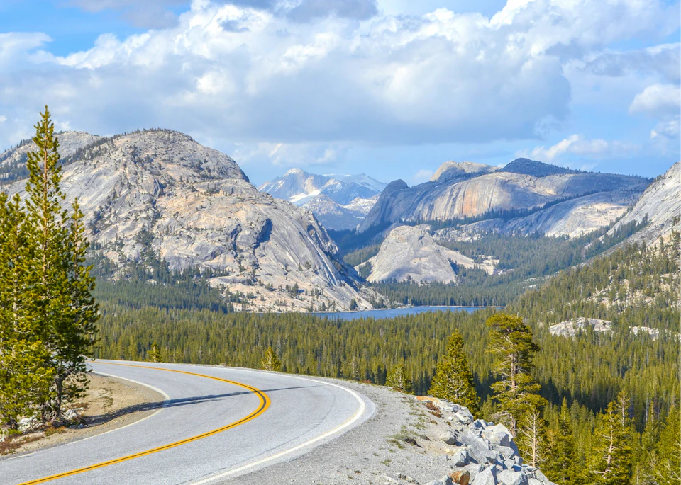 A winding road leading towards a scenic mountain range with a forest and a lake under a partly cloudy sky.