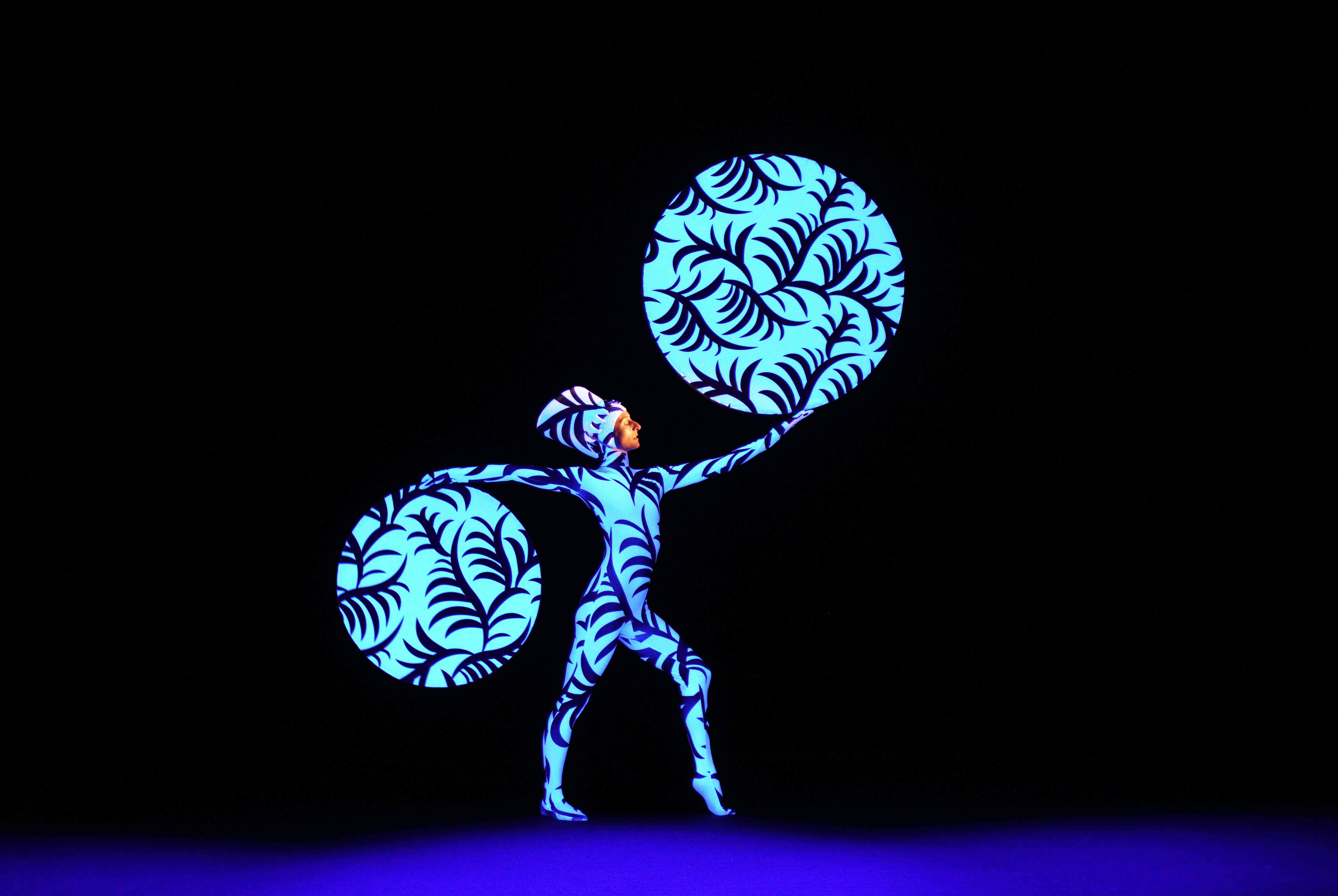 A performer in a blue and black patterned bodysuit holds two large, similarly patterned circular props on a dark stage.