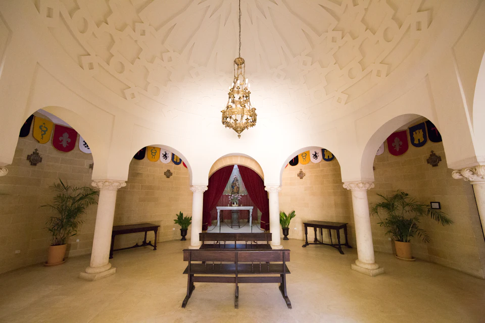 A small chapel with arched doorways, a chandelier, benches, and an altar with red curtains, potted plants, and decorative flags.