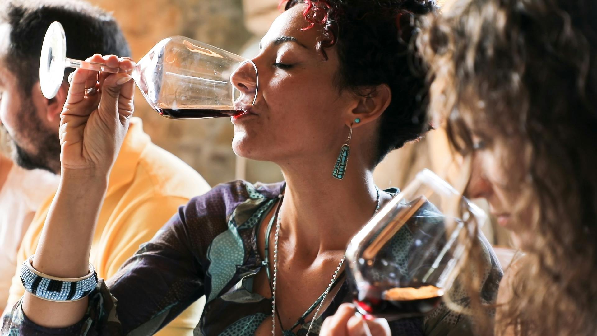 短いカーリーヘア、イヤリングを着用し、柄のトップスを着て、グラスから赤ワインを飲んでいる人、他の人と一緒に近くにいます。