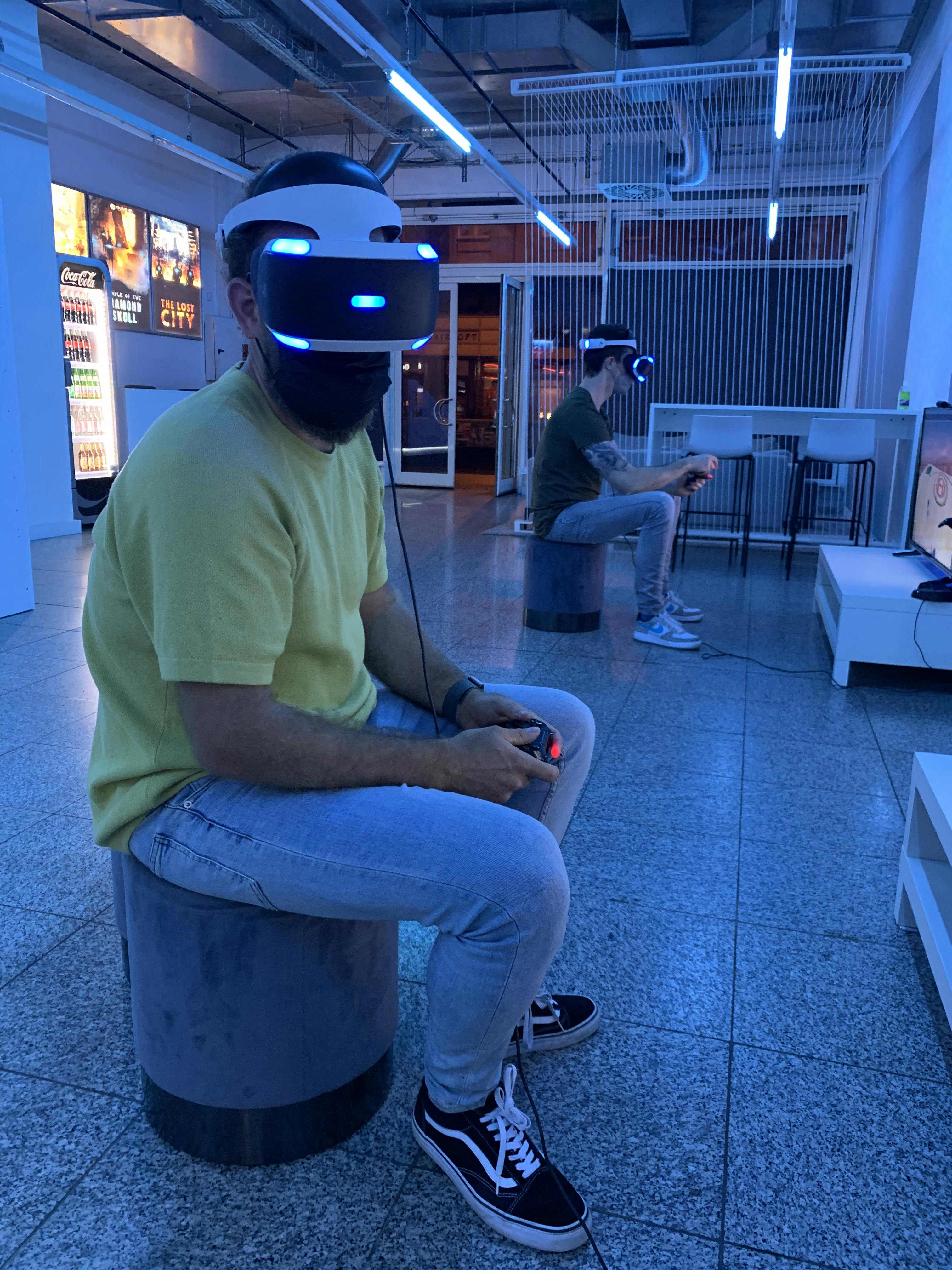 파란색 조명과 자동 판매기를 배경으로 방에 앉아 VR 헤드셋을 착용하고 컨트롤러를 들고 있는 두 사람.