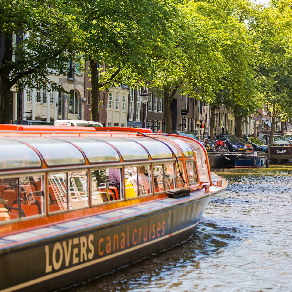 Canal boat with "LOVERS canal cruises" text on side, gliding through a tree-lined canal with parked cars and houses in the background.