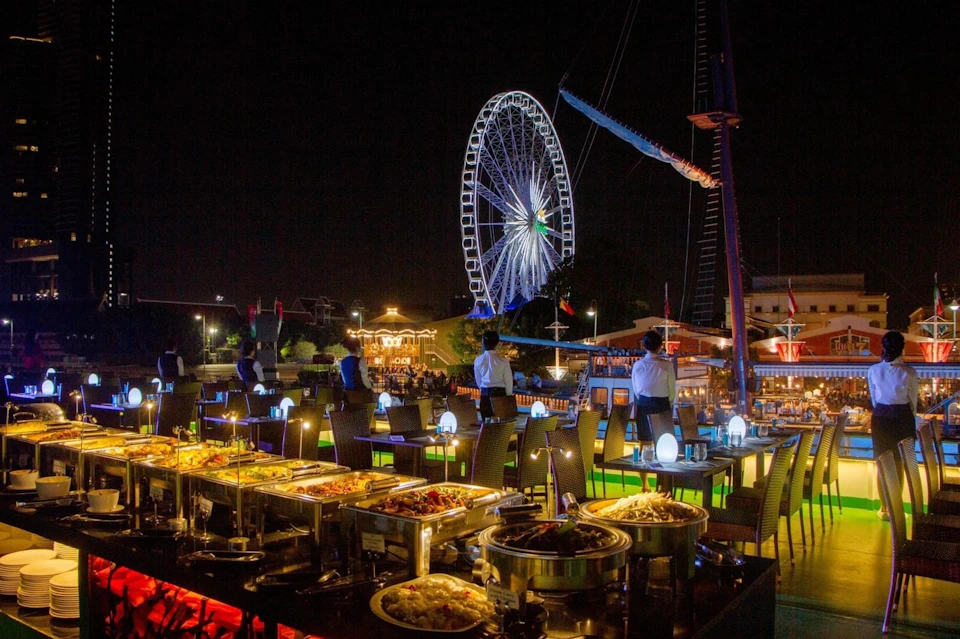 Nighttime outdoor dining area with buffet tables, lit decorations, and a large illuminated Ferris wheel in the background.