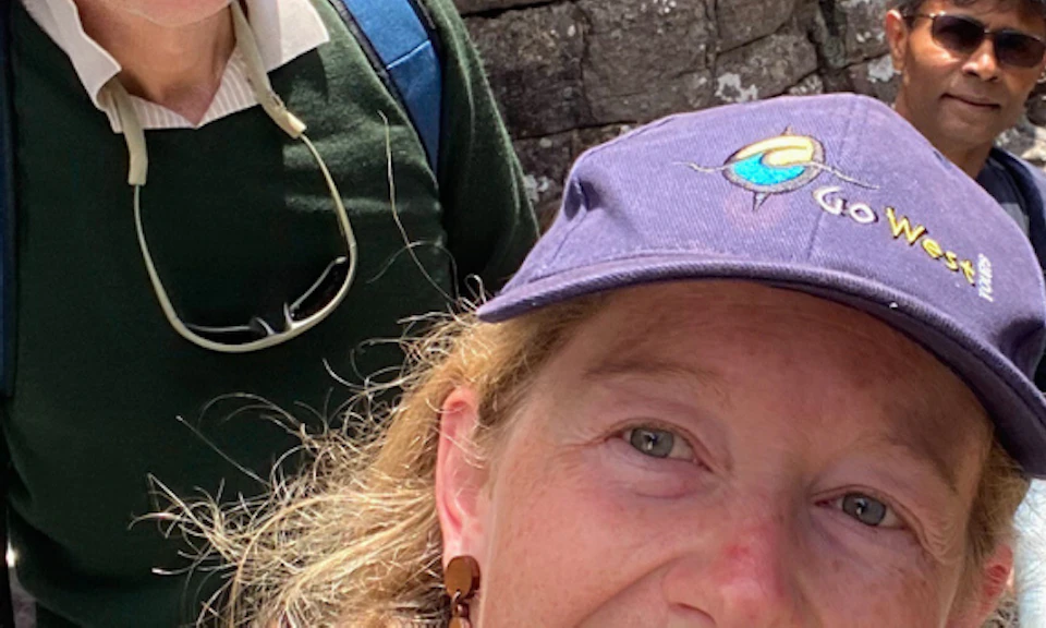 A group of people smiles on a rocky hiking trail. The woman in front takes a selfie, with others climbing stairs behind her.