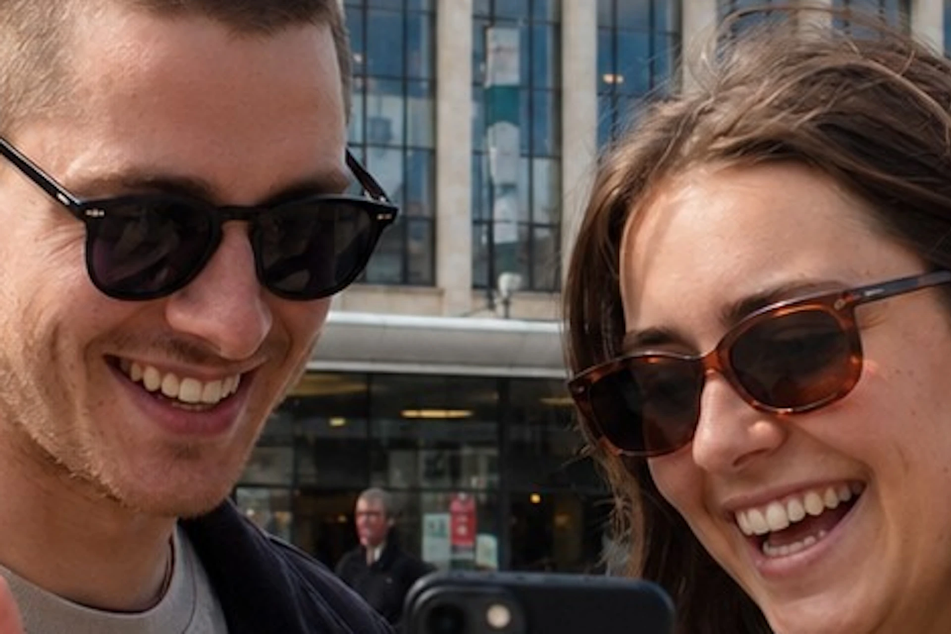 Two people outdoors, smiling while looking at a smartphone, with a modern building in the background.