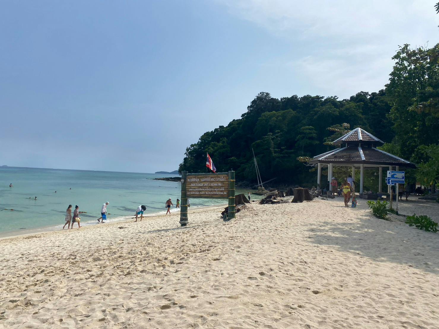 Scène de plage avec des gens marchant sur le sable, un panneau en bois, un pavillon et une colline boisée en arrière-plan.