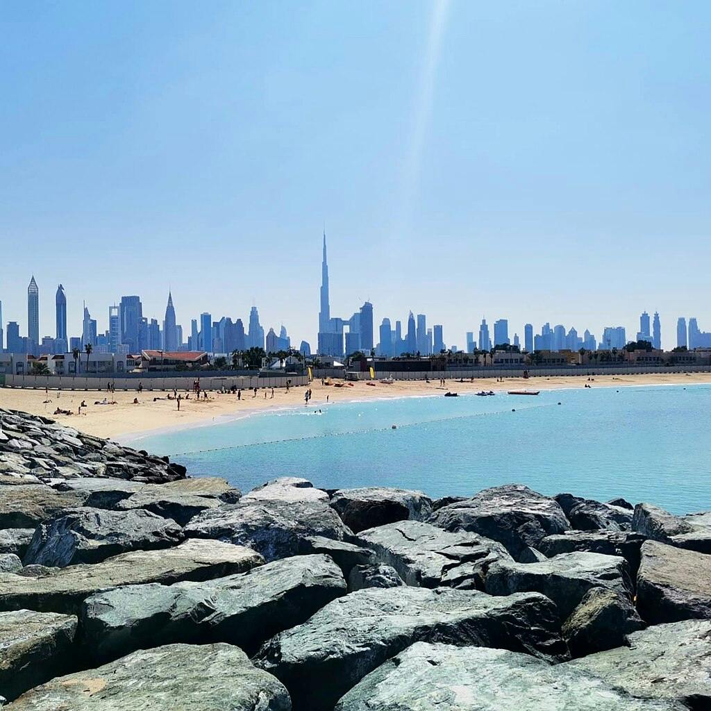 Costa rocosa en primer pla, una platja de sorra amb gent al mig i un skyline de la ciutat amb edificis alts al fons.