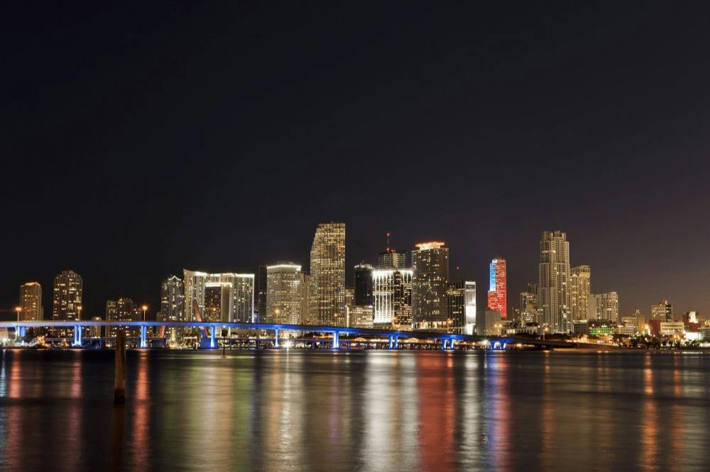 A city skyline at night with illuminated buildings, a lit-up bridge in the foreground, and reflections on the water below.