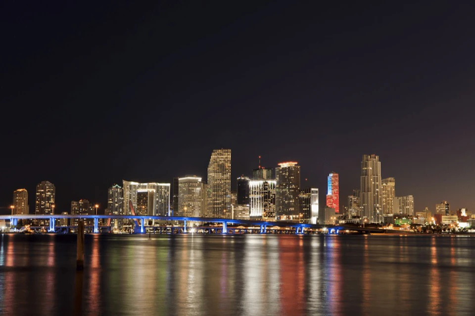 A city skyline at night with illuminated buildings, a lit-up bridge in the foreground, and reflections on the water below.
