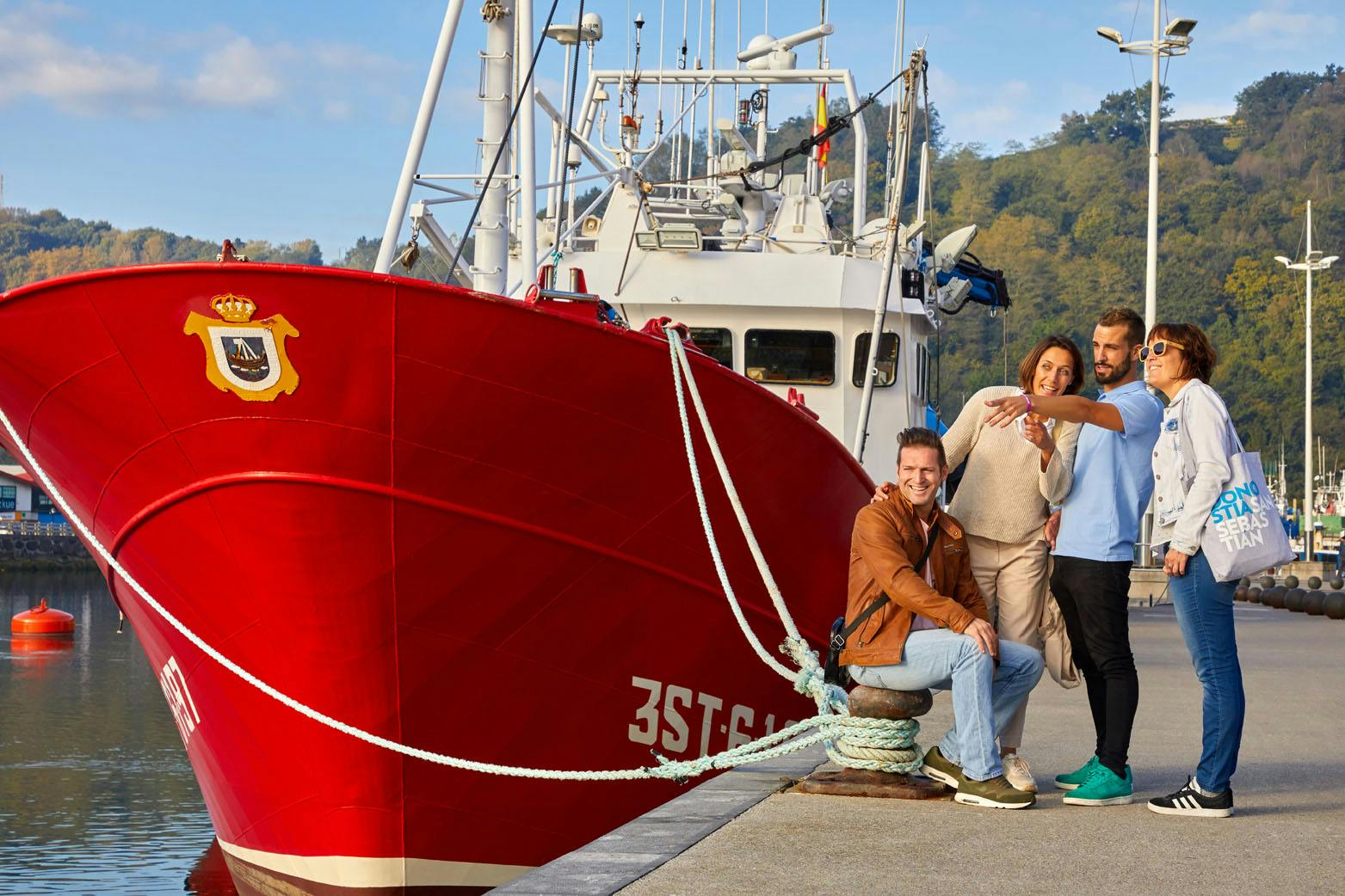 Four people smiling and posing for a photo by a docked red boat, with a wooded hillside in the background.
