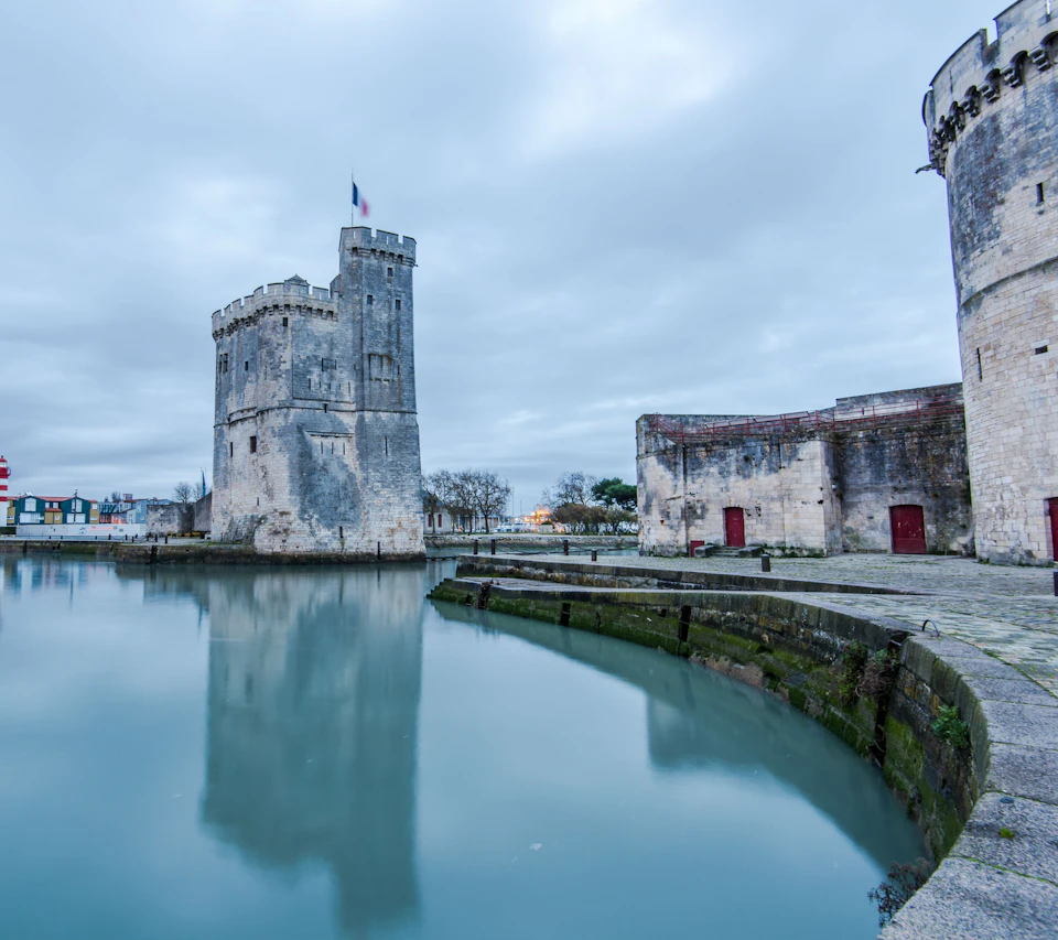 A tall, historic stone tower with a flag stands beside calm water, with additional stone structures and cloudy sky in the background.
