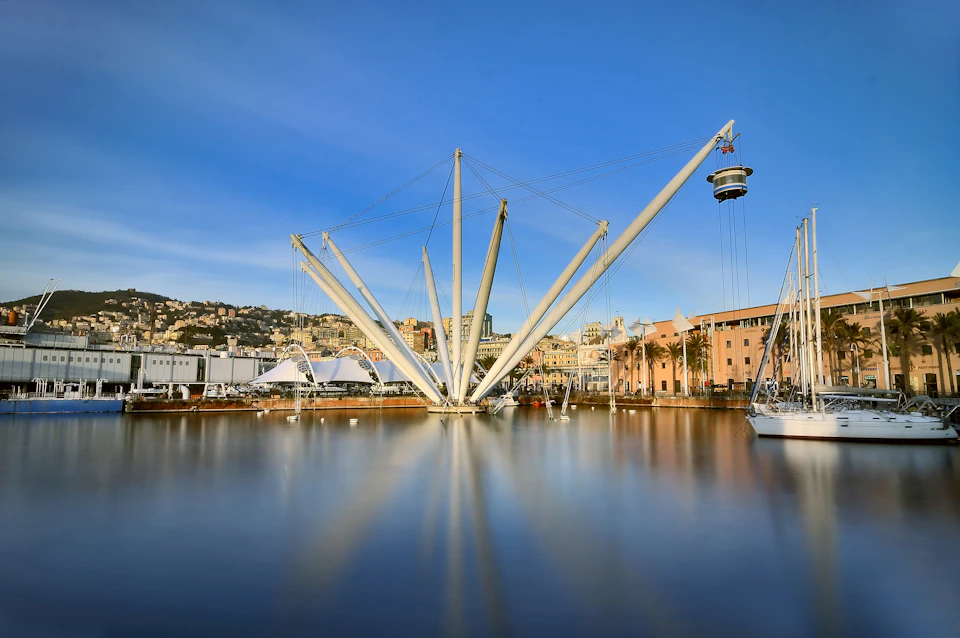 Modern harbor structure with steel arms and cables, surrounded by water and boats, with buildings and hills in the background.