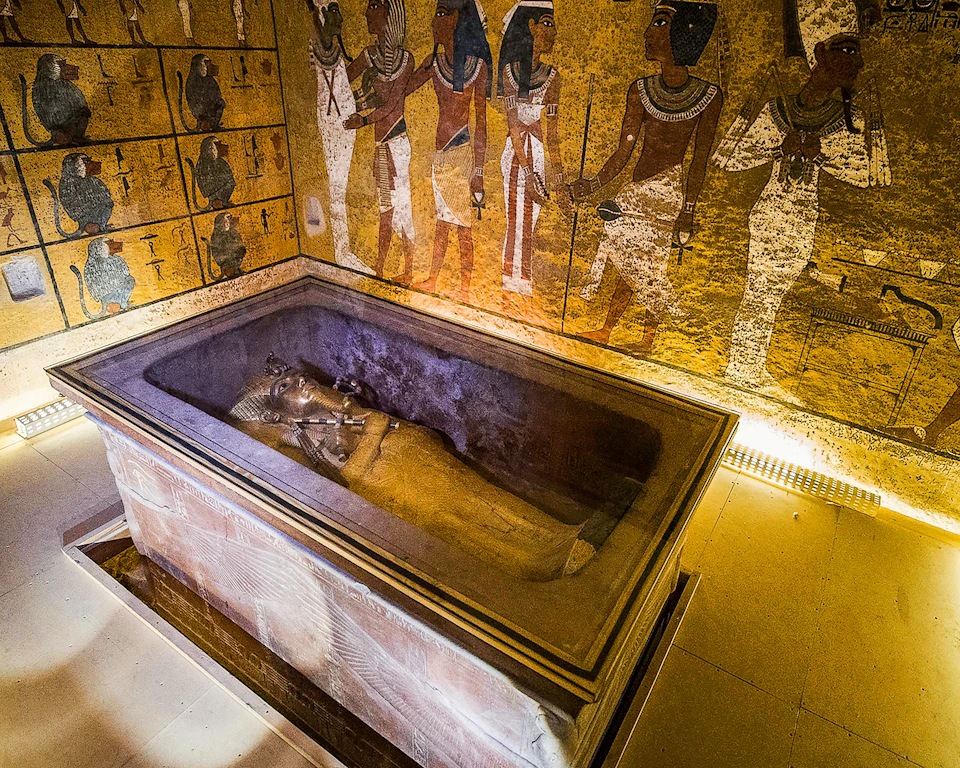 An ancient Egyptian sarcophagus in a decorated tomb, with hieroglyphic-covered walls depicting figures in traditional attire.
