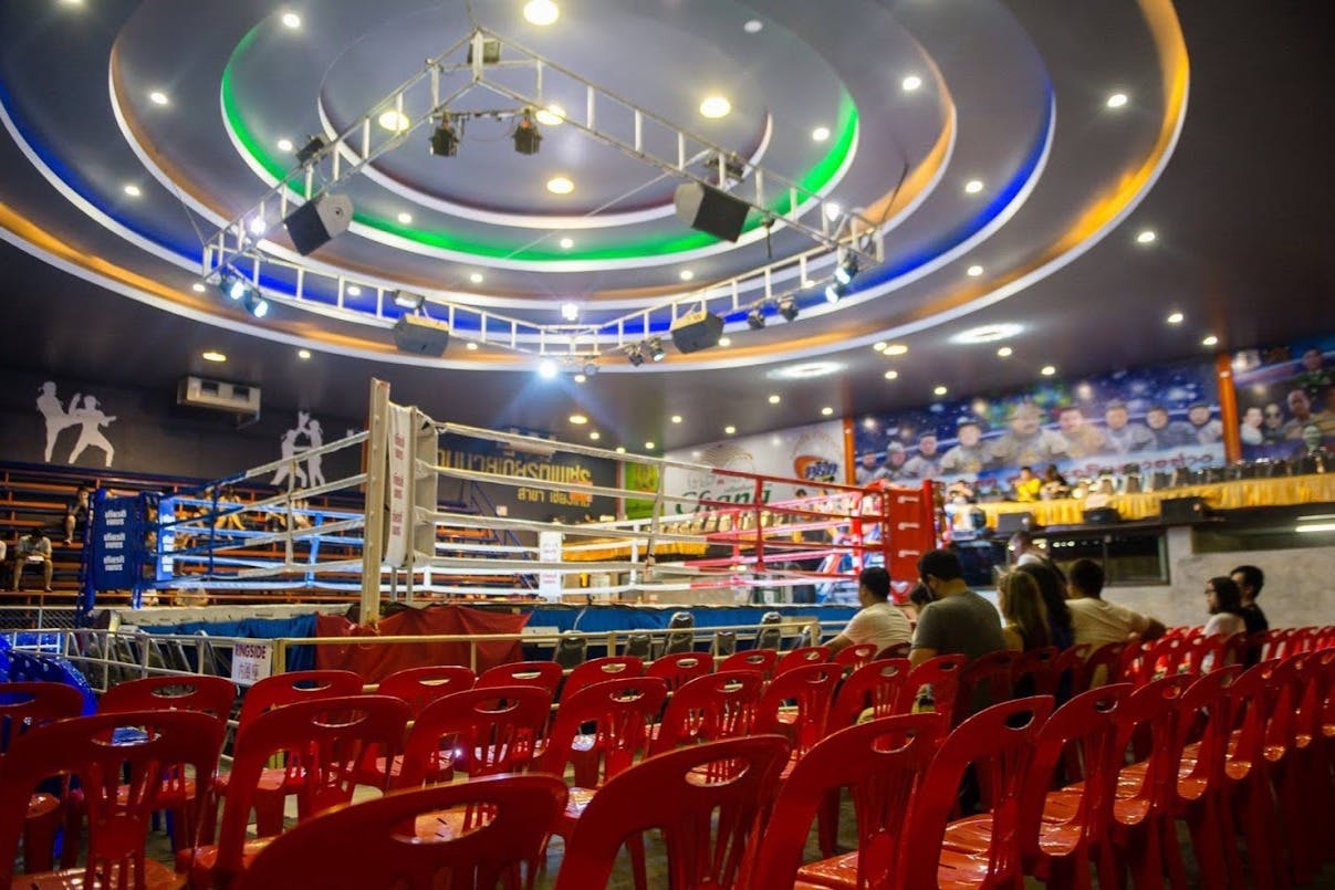 Un ring de boxe intérieur entouré de chaises rouges et de spectateurs, avec un éclairage coloré et un plafond décoré.
