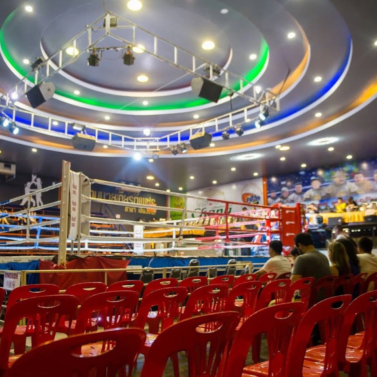 An indoor boxing ring surrounded by red chairs and spectators, with colorful lighting and a decorated ceiling.