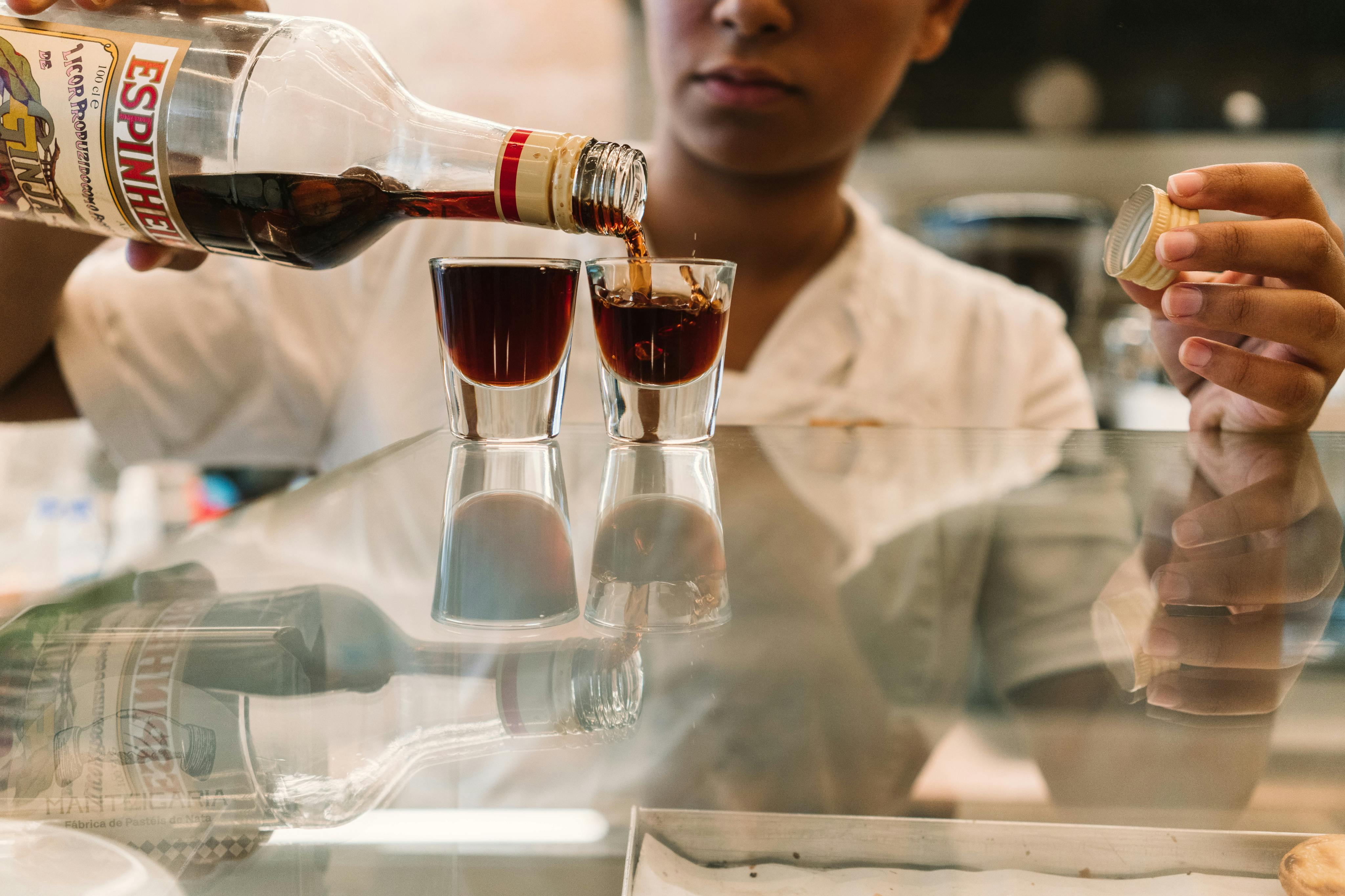 Een persoon giet donkere vloeistof in twee kleine glazen op een reflecterend oppervlak, terwijl hij een fles en een dop vasthoudt.