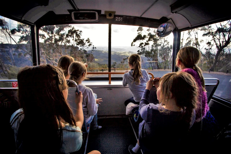 Children inside a bus looking out the large front window at a mountainous landscape with trees and distant hills.