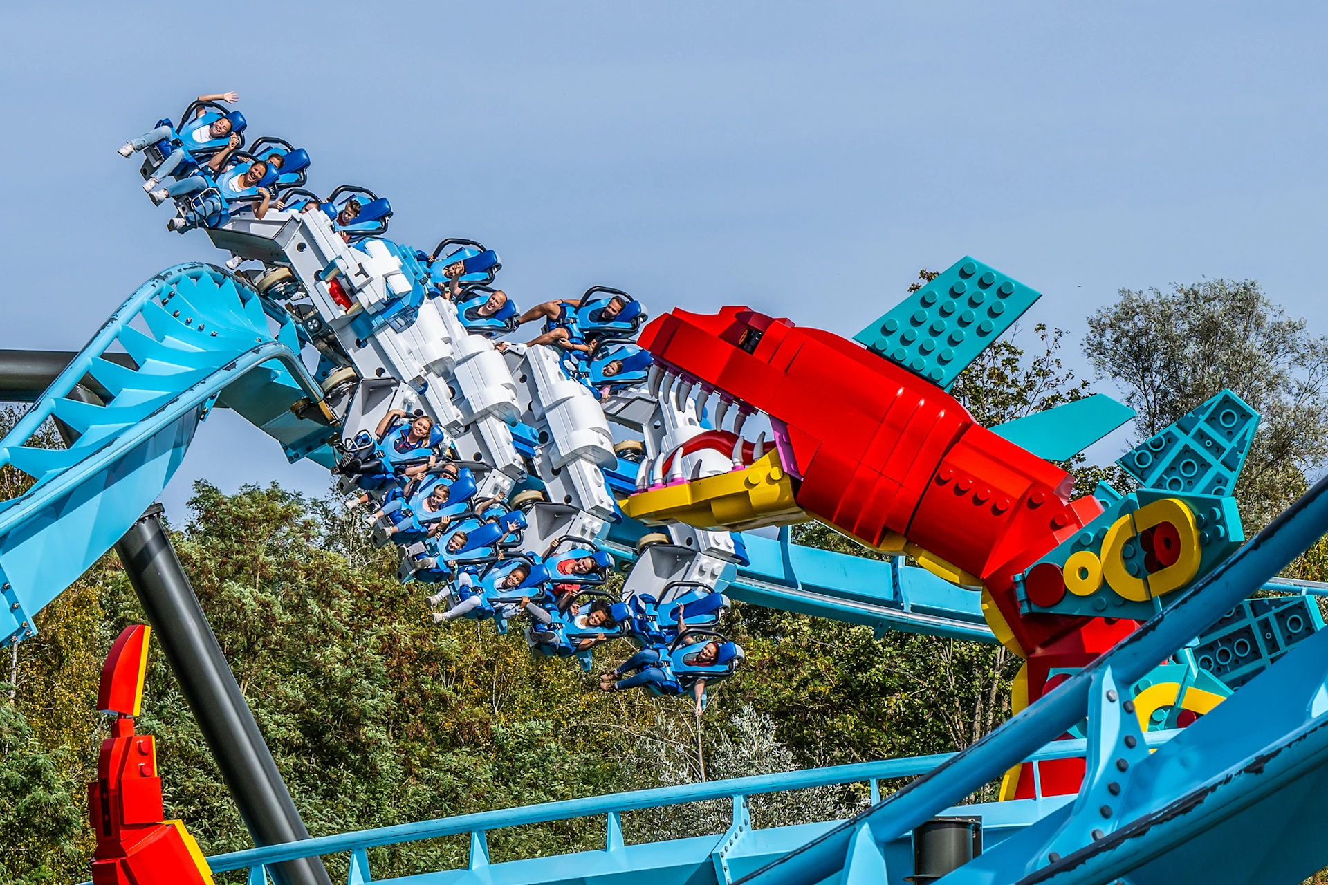 A rollercoaster with passengers passes by a colorful red and yellow LEGO-style shark figure, surrounded by trees.